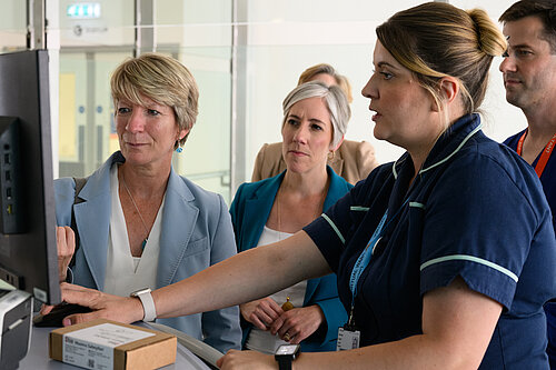 A nurse at Addenbrookes Hospital demonstrates something on a screen to Pippa Heylings, Lib Dem Parliamentary Candidate for South Cambs. Daisy Cooper MP,  the national Liberal Democrat Health and Social Care spokesperson, sits behind Pippa looking at the same screen.