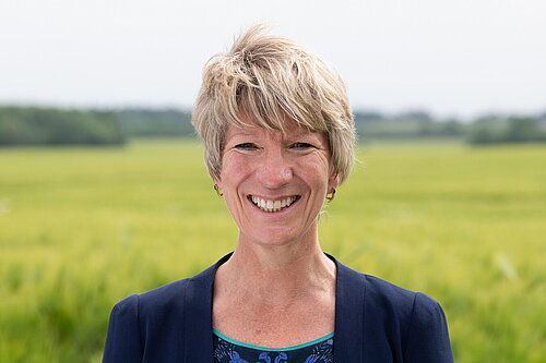 Head and shoulders photo of Pippa Heylings, Lib Dem Parliamentary candidate for South Cambs. Pippa is smiling at the camera, standing in front of a grassy field with trees in the distance.