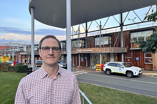 Ian Sollom standing across the road from the main entrance to Hinchingbrooke Hospital, 