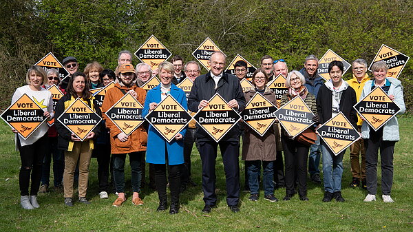 A group of about 20 Lib Dems from South Cambridgeshire standing in front of some trees, holding Lib Dem banners and cheering. Pippa Heylings parliamentary candidate for South Cambs is standing in the centre with Ed Davey, the party leader, standing next to her.