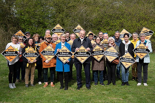 A group of about 20 Lib Dems from South Cambridgeshire standing in front of some trees, holding Lib Dem banners and cheering. Pippa Heylings parliamentary candidate for South Cambs is standing in the centre with Ed Davey, the party leader, standing next to her.